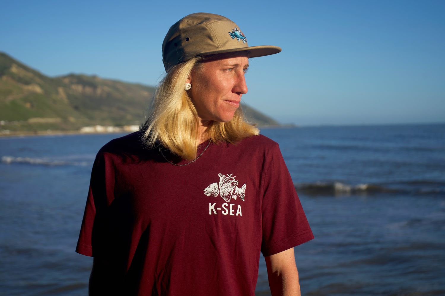Person wearing the Ocean Pulse maroon t-shirt design of a surreal fish with a human heart and a khaki cap with the K-Sea logos, looking to the side in a moody and slightly darker ocean scene background in Ventura, California.